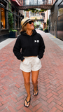 A woman walking down a brick alley wearing a black cropped hoodie with a white play/pause symbol on the chest, white drawstring shorts, a straw hat, and black flip-flops.