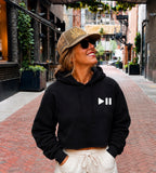 A woman wearing a black cropped hoodie with a white pause button graphic on the chest, cream drawstring pants, and a brown trucker hat with 'LOCAL SUMMER' printed on it, standing in a brick alleyway. Five local summer foam trucker hats in Olive, sunny, Jade, Sandy and coral with gold lettering, mesh backs, and adjustable snapback closures by token jersey.
